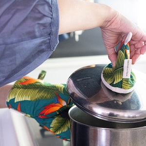 May include: A person wearing a green and orange pot holder with a tropical print is holding a green and white pot holder with a leaf design. The pot holder has a white tag that says "Handmade in Hawaii".