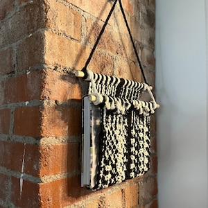 May include: A black and white macrame wall hanging with wooden dowels, hanging on a brick wall. The wall hanging is designed to hold books or magazines.