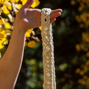 Puede incluir: Colgador de plantas de macramé color crema con un patrón de diamantes. El grueso cordón tejido se sostiene contra un fondo borroso de follaje amarillo y verde. El lazo superior es visible, listo para colgar.