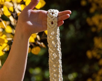 May include: Cream-colored macrame plant hanger with a diamond pattern. The thick, woven cord is held up against a blurred background of yellow and green foliage. The top loop is visible, ready for hanging.