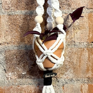 Puede incluir: Un macramé blanco para colgar plantas con una maceta de terracota y una planta de color morado oscuro. El macramé tiene cuentas de madera y una etiqueta negra que dice "Hand Made".