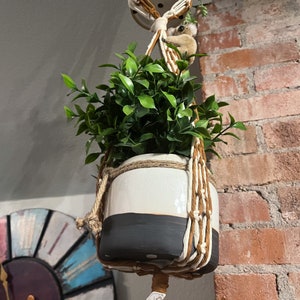 May include: A white and black planter with a macrame hanger, filled with green leafy plants. The planter is hanging from a brick wall.