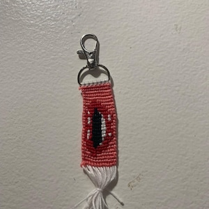 May include: A handmade keychain featuring a woven design with a red and white pattern, and a black eye in the center. The keychain has a silver clasp and a white tassel at the bottom.