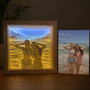 May include: A square wooden light box featuring a backlit layered paper cutout of two people posing on a beach, displayed next to the original color photograph showing two people in swimsuits standing on the sand by the ocean waves.