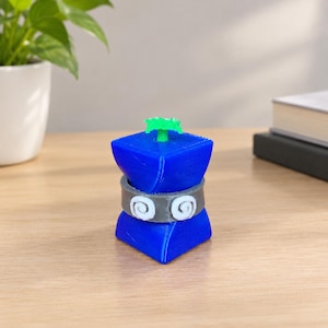 May include: A small, blue, 3D-printed cube-shaped figurine with a gray band around the middle featuring two white spiral designs. A bright green, textured, star-shaped sprout protrudes from the top center of the blue cube structure.