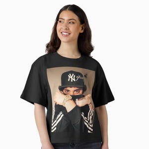 May include: Black t-shirt featuring a portrait of a person wearing a black baseball cap with a white "NY" logo and a black jacket with white stripes. The person is wearing a gold watch and rings.