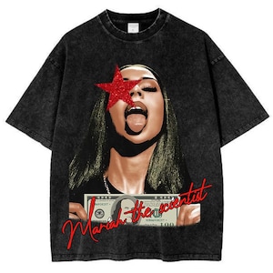 May include: Black graphic t-shirt featuring a woman with a red star over one eye and her tongue out. The text "Mariah the scientist" is written in red cursive across the bottom of the image. The shirt is made of a dark, washed fabric.