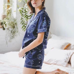 May include: A navy blue pajama set featuring a short-sleeved button-down shirt and matching shorts. The fabric is decorated with a repeating white and light blue eye pattern, accented with small star shapes and white piping along the edges.