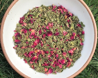 May include: A white bowl filled with a blend of dried herbs and rose petals. The herbs are a mix of green and brown, and the rose petals are a vibrant pink.