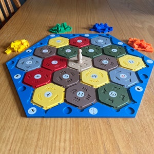 May include: A hexagonal game board with blue edges and a variety of coloured hexagonal tiles. The tiles are numbered 2 through 12 and have different symbols on them. A wooden game piece is in the centre of the board.