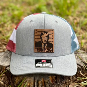 May include: Gray trucker hat with a leather patch featuring a portrait and the text "TRUMP 2024." The hat has a curved brim and a mesh back with red, white, and blue accents. The Richardson brand tag is visible.