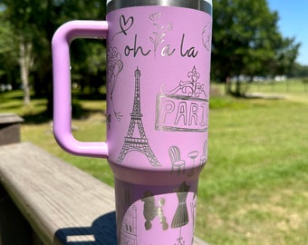 May include: A lavender travel mug with a handle, featuring a silver design of Parisian landmarks, including the Eiffel Tower, a poodle, and the words "oh la la", "Paris", and "Bonjour".