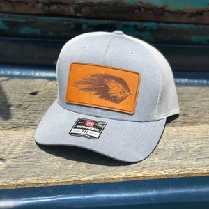 May include: A gray and white mesh trucker hat with a brown leather patch featuring a fly fishing lure. The patch is stitched onto the front of the hat. The hat has a Richardson 112 adjustable strap.