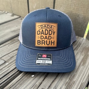May include: A navy blue and gray trucker hat with a brown leather patch that says "*DADA* DADDY >DAD< BRUH". The hat is adjustable and has the Richardson logo on the back.