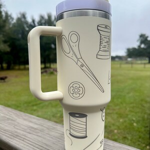 Op de afbeelding: Een witte reistumbler met een zwarte handgreep en een ontwerp van naai-accessoires, waaronder een schaar, spoelen garen, knopen en een speldenknop.