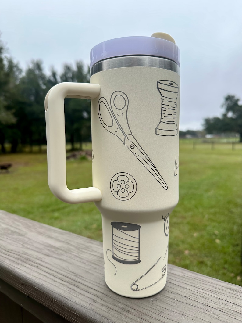 Op de afbeelding: Een witte reistumbler met een zwarte handgreep en een ontwerp van naai-accessoires, waaronder een schaar, spoelen garen, knopen en een speldenknop.