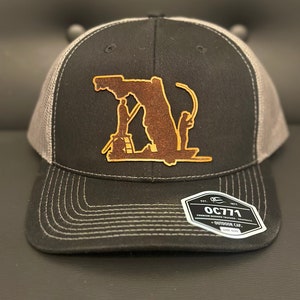 May include: Black and gray trucker hat with a brown leather patch depicting the Florida state outline with a fishing scene. The hat has a curved brim and a white stitched design. The tag reads "OC771".