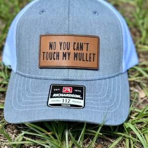 May include: A light blue trucker hat with a brown leather patch that says "NO YOU CAN'T TOUCH MY MULLET". The hat has a white mesh back and a Richardson logo on the front.