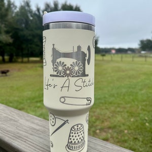Op de afbeelding: Een witte roestvrijstalen tumbler met een lavendelkleurig deksel. De tumbler heeft een naaimachine-ontwerp met de tekst "Life's A Stitch".