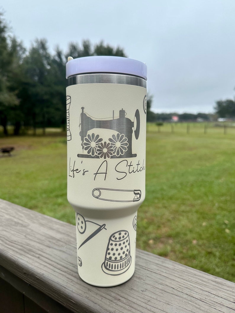 Op de afbeelding: Een witte roestvrijstalen tumbler met een lavendelkleurig deksel. De tumbler heeft een naaimachine-ontwerp met de tekst "Life's A Stitch".