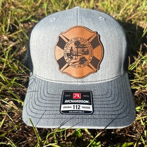 May include: Gray and black trucker hat with a brown leather patch featuring the Great Seal of the State of Florida. The patch has the text "Great Seal of the State of Florida" and "In God We Trust". The hat has a black mesh back and a Richardson 112 label.