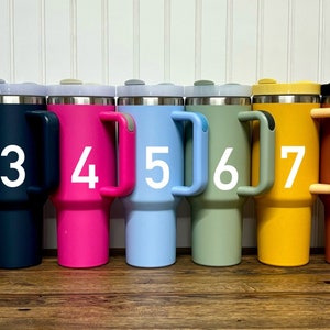 Op de afbeelding: Negen reistassen van roestvrij staal met handvatten. De bekers zijn in verschillende kleuren, waaronder blauw, grijs, marineblauw, roze, lichtblauw, groen, geel, oranje en wit. Elke beker heeft een nummer van 1 tot 9 gedrukt op de zijkant.