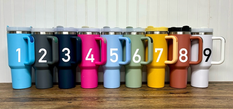 Op de afbeelding: Negen reistassen van roestvrij staal met handvatten. De bekers zijn in verschillende kleuren, waaronder blauw, grijs, marineblauw, roze, lichtblauw, groen, geel, oranje en wit. Elke beker heeft een nummer van 1 tot 9 gedrukt op de zijkant.