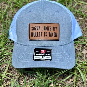 May include: A light blue trucker hat with a white mesh back. The hat has a brown leather patch with the text "SORRY LADIES MY MULLET IS TAKEN". The hat also has a red and black label with the text "RICHARDSON ORIGINAL 112 TRUCKER YOUTH".