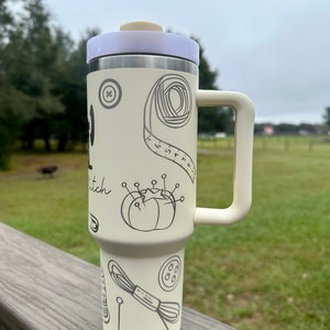 Op de afbeelding: Een witte roestvrijstalen reistumbler met een zwarte handgreep. De tumbler heeft een ontwerp met naai-gerelateerde items, waaronder een knoop, een spoel garen, een meetlint en een naaldkussen. De tekst "Stitch" is zichtbaar op de tumbler.