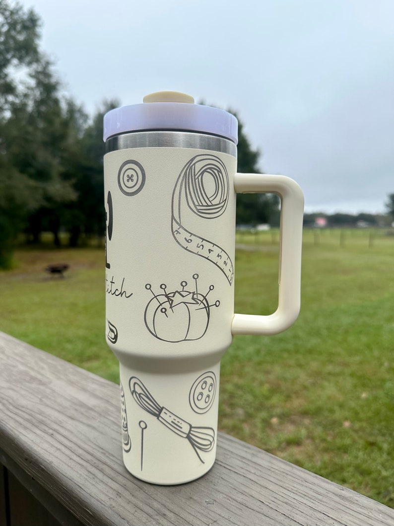 Op de afbeelding: Een witte roestvrijstalen reistumbler met een zwarte handgreep. De tumbler heeft een ontwerp met naai-gerelateerde items, waaronder een knoop, een spoel garen, een meetlint en een naaldkussen. De tekst "Stitch" is zichtbaar op de tumbler.