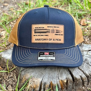 May include: A navy blue and brown trucker hat with a brown leather patch that says "Anatomy of a Pew" with a diagram of a bullet and the words "Bang Button", "Magical Fire Dust", "Metal Holding Thing", and "Freedom Seed". The hat is made by Richardson and is adjustable.