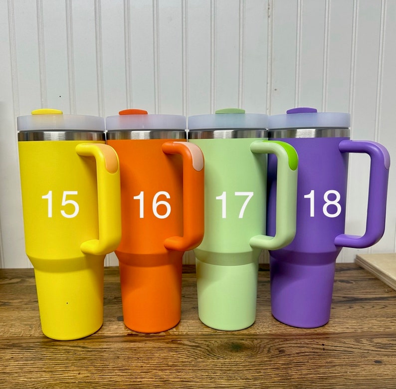 Op de afbeelding: Vier roestvrijstalen reistassen met handvatten. De bekers zijn geel, oranje, groen en paars. De bekers zijn genummerd 15, 16, 17 en 18.