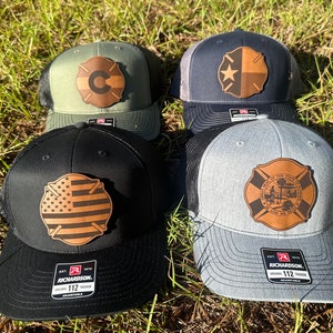 May include: Four different trucker hats with leather patches. The patches feature a fire department symbol with different state flags or emblems. The hats are in various colors, including green, black, navy blue, and gray.