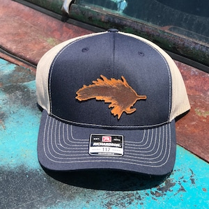 May include: A navy blue and beige trucker hat with a brown leather patch shaped like a leaf. The hat has a mesh back and an adjustable strap with the text "Richardson 112" on it.