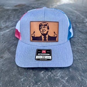 May include: A blue and red trucker hat with a brown leather patch featuring a black and white image of Donald Trump giving the middle finger. The patch has the text "EST. 1970 RICHARDSON. ORIGINAL 112 TRUCKER ADJUSTABLE".