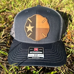 May include: A black and gray trucker hat with a brown leather patch featuring a star and a fire department symbol. The hat has an adjustable strap and a label that reads "Richardson. Est. 1970 Original 112 Trucker Adjustable".