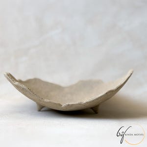 May include: A handcrafted, beige ceramic bowl with an irregular, organic form. The bowl features a textured finish and rests on three small feet. The design evokes a natural aesthetic, suitable for displaying small objects or as a decorative item.
