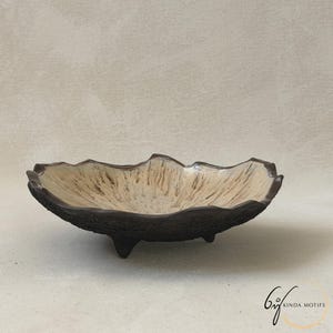 May include: A shallow ceramic bowl with a dark brown, textured exterior and a cream-colored interior featuring radiating brown streaks. The bowl has an irregular, wavy rim and stands on three small, dark brown feet. The text KINDA MOTIFS is visible.