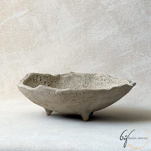 Peut inclure: Un petit bol en céramique gris clair texturé, avec un bord irrégulier unique. Le bol repose sur trois petits pieds arrondis. La surface a un aspect poreux et vieilli, suggérant une origine artisanale. Le bol est probablement utilisé à des fins décoratives.