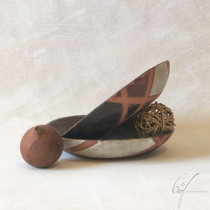 May include: Two stacked ceramic bowls featuring a geometric pattern of dark brown, terracotta, and off-white segments. A small, round, matte terracotta sphere sits to the left, and a woven rattan sphere rests inside the lower bowl.