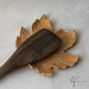 May include: A wooden spatula rests on a ceramic leaf-shaped spoon rest. The spoon rest is a warm, golden-yellow colour with detailed leaf vein patterns. The spatula is a dark brown colour.