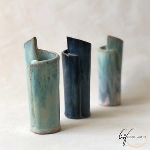 May include: Three ceramic vases in shades of blue and teal. Each vase has a unique, asymmetrical design with a curved opening. The vases have a textured, glazed finish, and are arranged in a row, showcasing their artistic form.