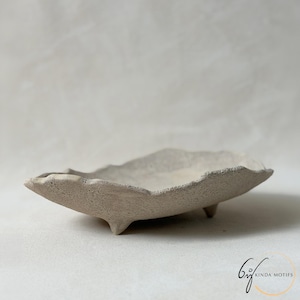 May include: A shallow, oval-shaped ceramic dish with a rough, textured, light grey surface and irregular, wavy edges. The dish is supported by three small, short feet and features the text KINDA MOTIFS in the bottom right corner.