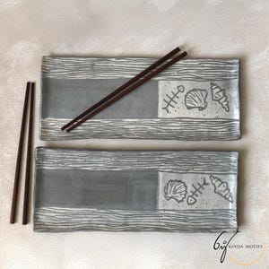 May include: Two rectangular grey ceramic sushi plates with a wood grain border and a grey centre. Each plate has a square section with seashell and fishbone designs. Two pairs of brown chopsticks are included.