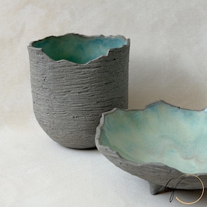 May include: A cylindrical ceramic cup and a shallow bowl with irregular, jagged rims. Both pieces feature a dark gray, heavily textured exterior and a smooth, vibrant turquoise blue interior glaze. The bowl is supported by small feet.