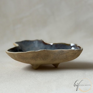 May include: A handmade, ceramic serving dish with a unique, leaf-like shape. The dish features a textured, tan exterior and a dark blue interior. It rests on three small feet and is ideal for serving snacks or small dishes.