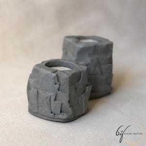 May include: Two grey, rock-shaped candle holders. The holders have a textured, rough surface and a cylindrical cavity to hold candles. The candle holders are in front of a neutral background. The logo "6if kinda motifs" is in the bottom right corner.
