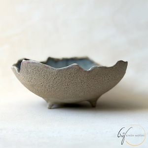 May include: A handmade ceramic bowl with a textured surface and a scalloped edge. The bowl is a neutral beige colour with a blue interior and stands on three small feet. Suitable for serving or as a decorative item.