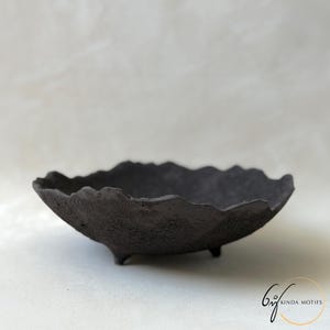 Peut inclure: Un bol noir charbon avec une surface rugueuse et texturée et un bord irrégulier et ondulé. Le bol est soutenu par trois petits pieds courts à la base. Dans le coin inférieur droit, il y a un logo avec le texte KINDA MOTIFS.