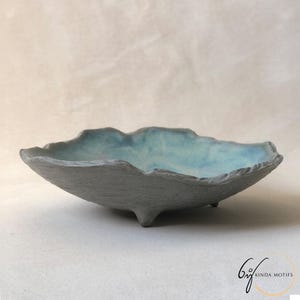May include: A handmade, grey ceramic bowl with an irregular, organic form. The interior has a light blue glaze, contrasting with the textured exterior. The bowl is supported by three small feet, enhancing its artistic design.